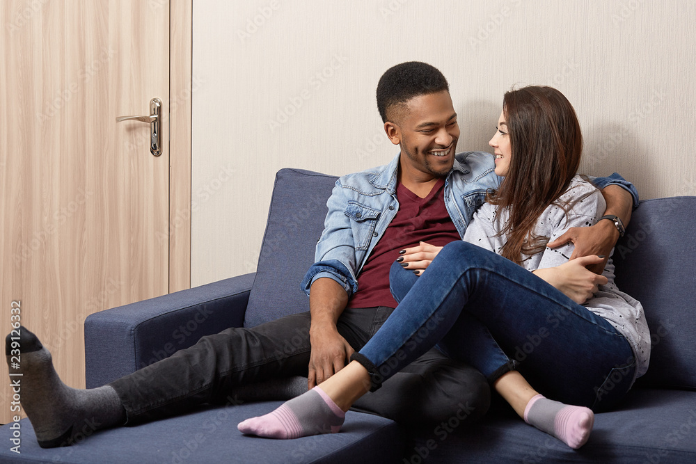 Obraz premium Affectionate mixed race couple in love look happily, have warm hug, dressed casually, pose at sofa in modern apartment, smile gladfully, speak about future, have good relationships. Multiethnic people