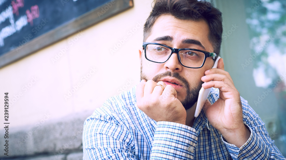 Young man in plaid shirt and eyeglasses has phone conversation, sitting at cafe. Young man in plaid shirt and eyeglasses has phone conversation, sitting at cafe.
