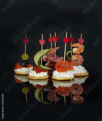 Assorted canapes of fresh goat cheese, salmon, salmon, red caviar, prosciutto on skewers with hearts on a dark background. On a Birthday, corporate or Valentine's day. Free space for text