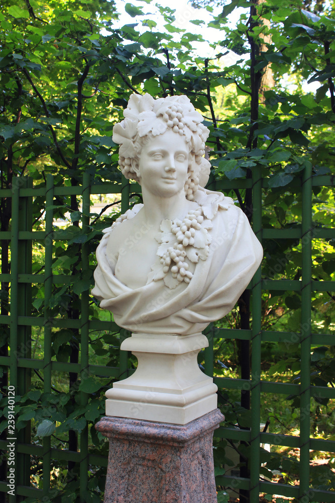 Fototapeta premium Antique Sculpture of Woman Head and Bust Representing Autumn in Summer Gardens. Historical White Statue Outdoors on Green Leaves Background at the City Park