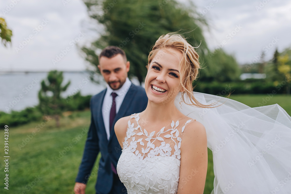 Blonde and smiling bride and bearded groom walk, holding hands, in the park with green grass and willows. Wedding portrait of beautiful newlyweds. Wedding photography. Blonde and smiling bride and bearded groom walk, holding hands, in the park with green grass and willows. Wedding portrait of beautiful newlyweds. Wedding photography.
