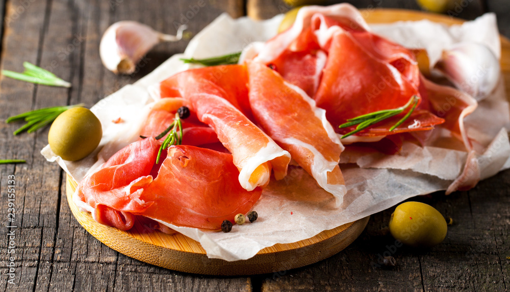 Closeup of thin slices of prosciutto with mixed olives and paprika on wooden cutting board