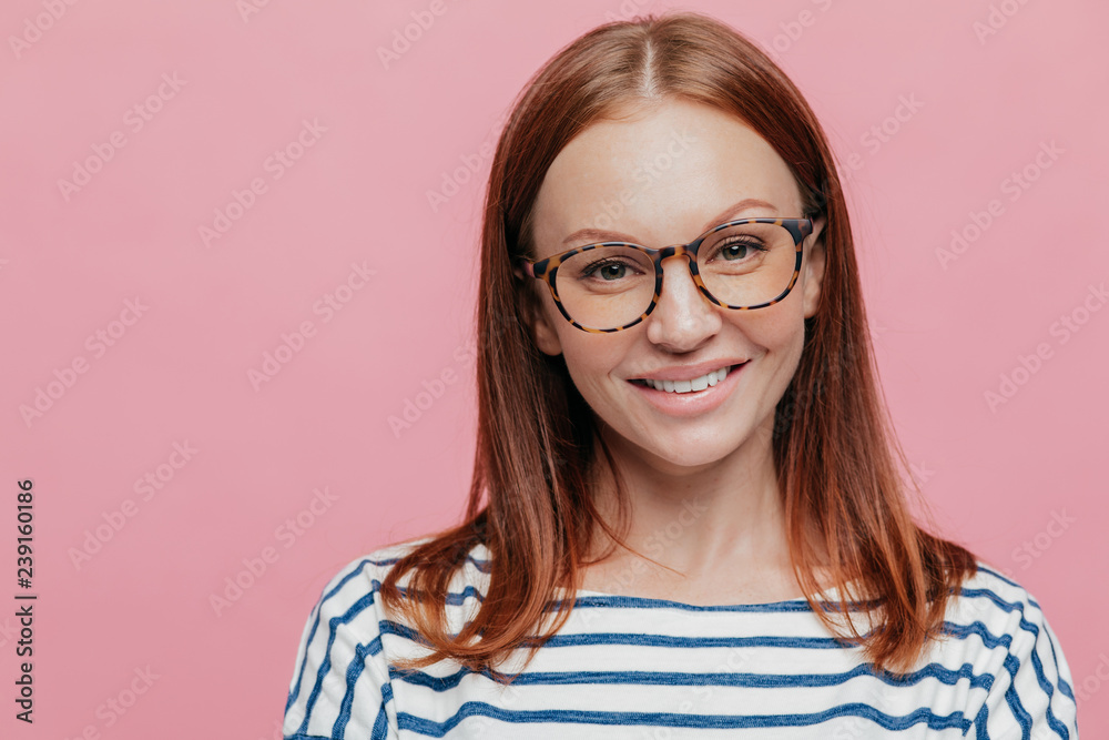 Close up shot of adorable young female student wears optical glasses, has tender smile, rejoices holidays wears striped clothes, models over pink background with copy space for your information.