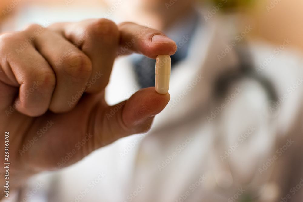 The white capsule in the hands of the doctor close up and the silhouette of a doctor in medical uniform in the background in soft focus. The white capsule in the hands of the doctor close up and the silhouette of a doctor in medical uniform in the background in soft focus.