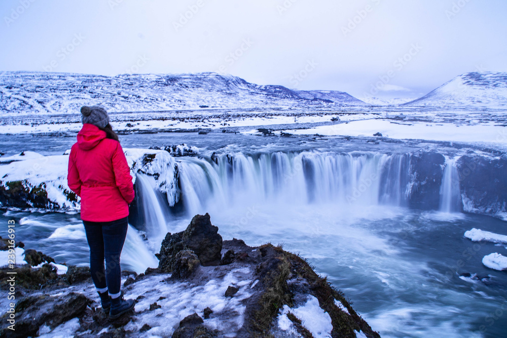 Scenic landscape view of tourist popular attraction Godafoss waterfall in northern Iceland in winter time. Young tourist woman overlooking falling water and snow covered mountains on background. 