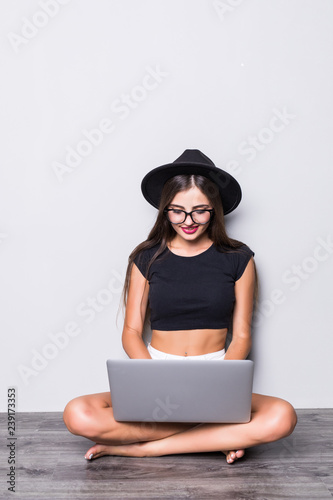 Smiling woman working on laptop computer while sitting on the floor with legs crossed isolated over gray background
