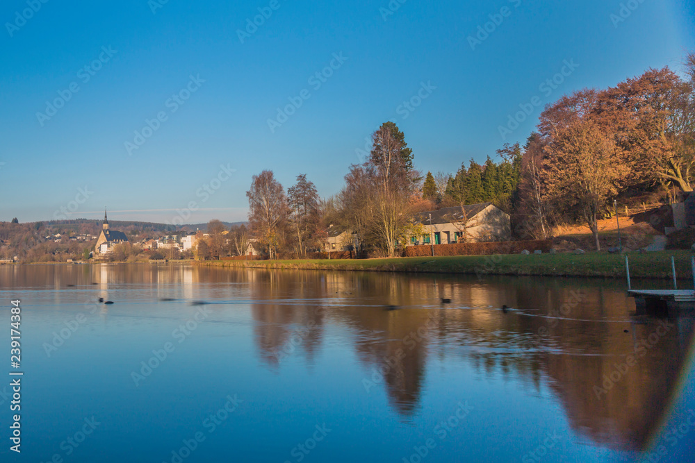 Landscape of Doyards lake with first rays of sunlight illuminating village of Vielsalm against clear blue sky in background, ducks swimming on calm waters on autumnal day in the Belgian Ardennes Landscape of Doyards lake with first rays of sunlight illuminating village of Vielsalm against clear blue sky in background, ducks swimming on calm waters on autumnal day in the Belgian Ardennes