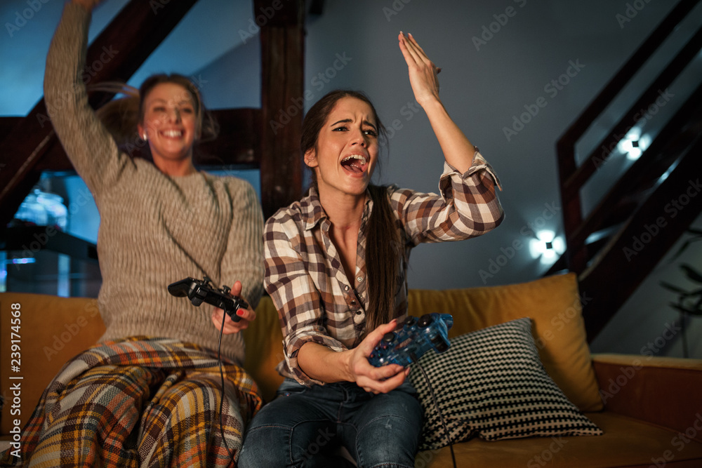 Two female best friends sitting at home on pleasant evening and playing games on console.One  friend lose challenge.