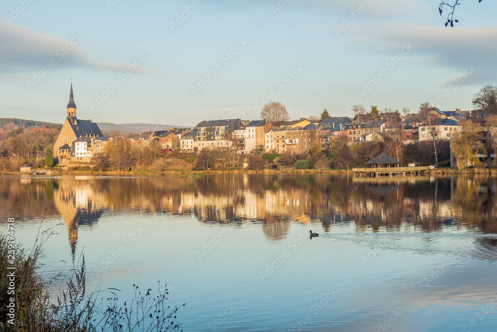 beautiful view of the Doyards lake with reflection of the village of Vielsalm and its St Gengoul church with a blue sky on a wonderful cold winter afternoon in the Belgian Ardennes