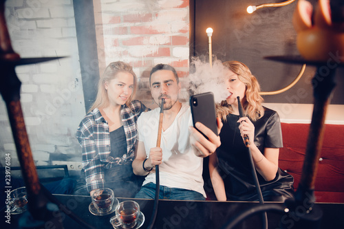 Guy smoking in hookah bar, take selfie, two beautiful blonde girls look at camera