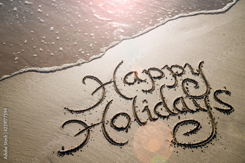 Happy Holidays message handwritten in smooth sand with an oncoming wave in the lens flare of the tropical sun