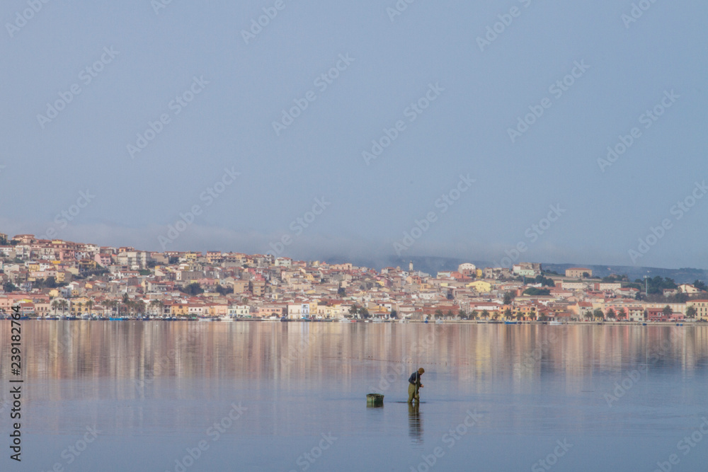 A fisherman at work bending in the flat sea water that reflects the houses of Sant'Antioco in Sardinia