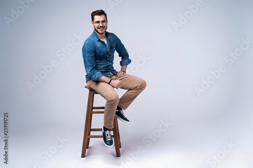 Full length portrait of an attractive young man in jeans shirt sitting on the chair over grey background.