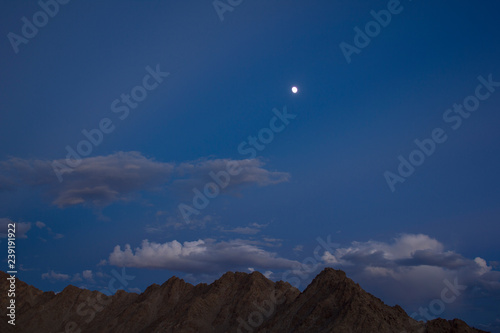 a ridge of gray brown desert mountains under a dark blue evening sky with gray clouds and a full moon with stars