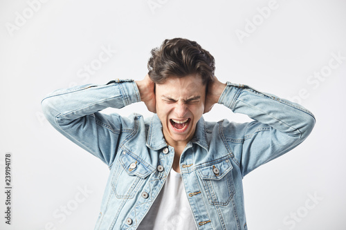 Dark-haired guy dressed in a white t-shirt and a denim jacket covers his ears with his hands and shouts on the white background in the studio