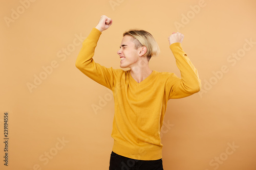 Joyful young blond guy dressed in yellow sweater is posing in the studio on the beige background
