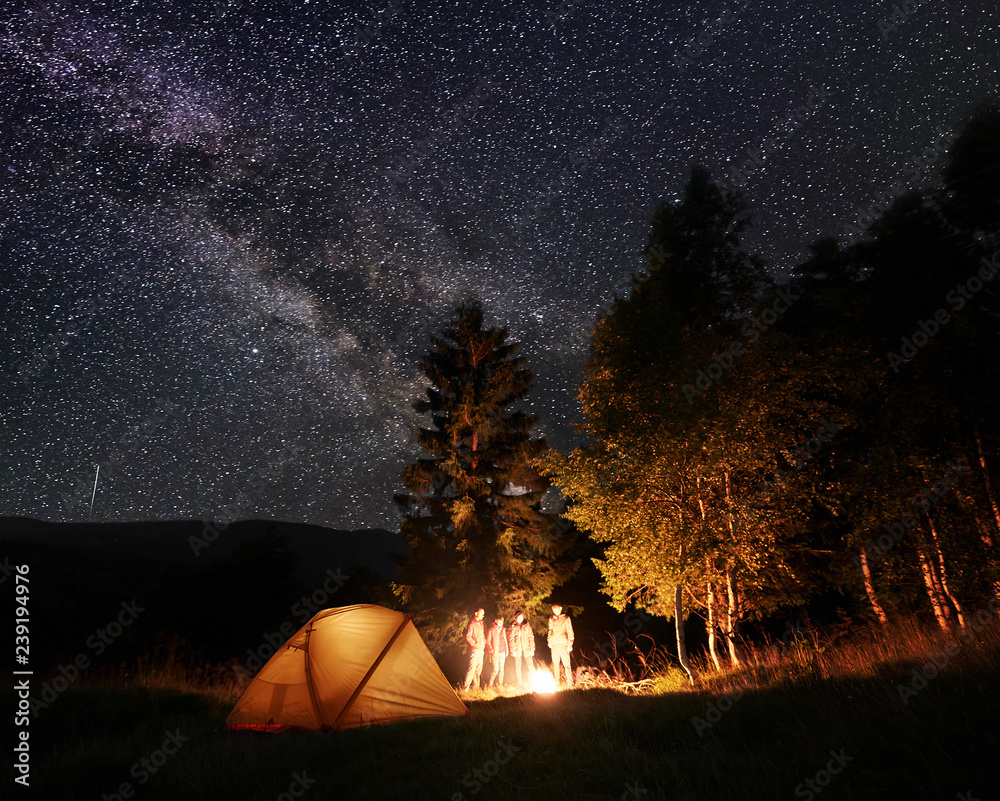 Plakat Group of tourists at night camping near the forest at the illuminated tent, looking at the burning fire under incredible beautiful starry sky and Milky way on the background of mountains and hills.