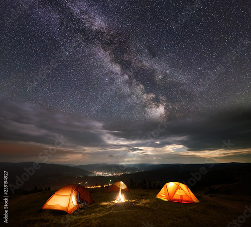 Night camping. Three illuminated orange tents in the mountains at night under starry sky, Milky way and clouds on the background of the luminous town in the valley. Between the tents lit a bonfire