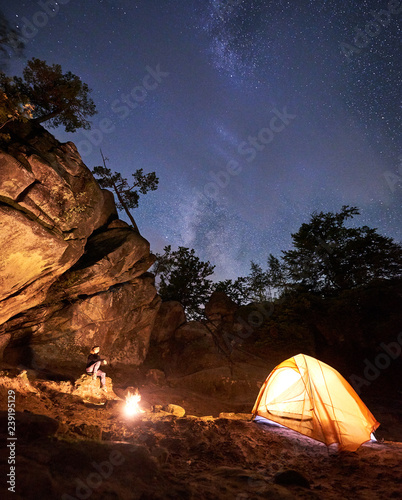 Campsite night amid huge steep rock formations. Attractive woman sitting on boulder watching bonfire in front of tourist tent on blue starry sky background. Tourism  sport and camping concept.