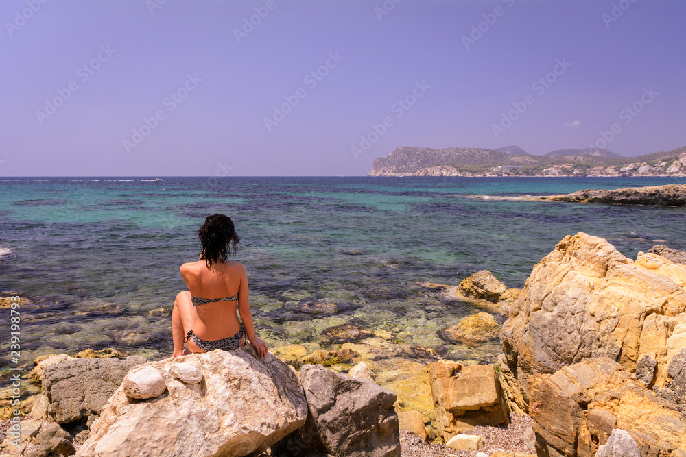 Young beautiful girl in a bikini sits on the rocks on the shores of the Mediterranean Sea. Mallorca, Balearic Islands, Spain