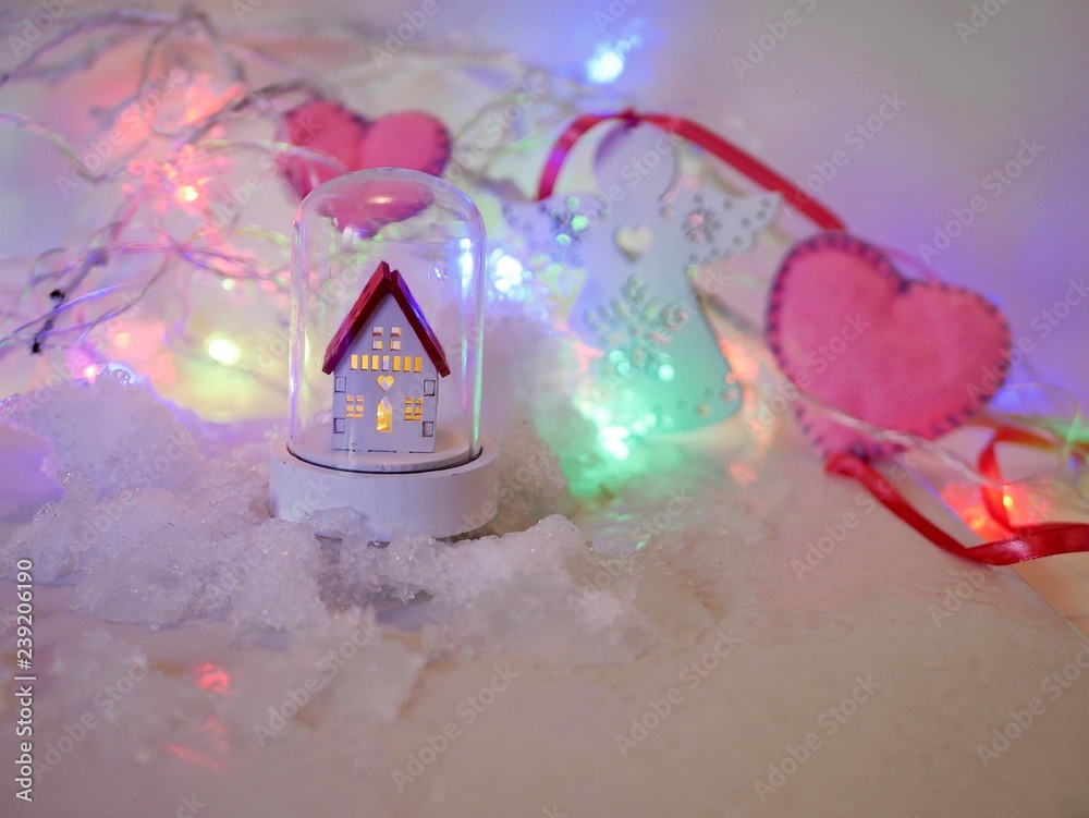  White toy house with a red roof, pink felt hearts against a background of lighted illumination on the snow, concept of winter seasonal holidays