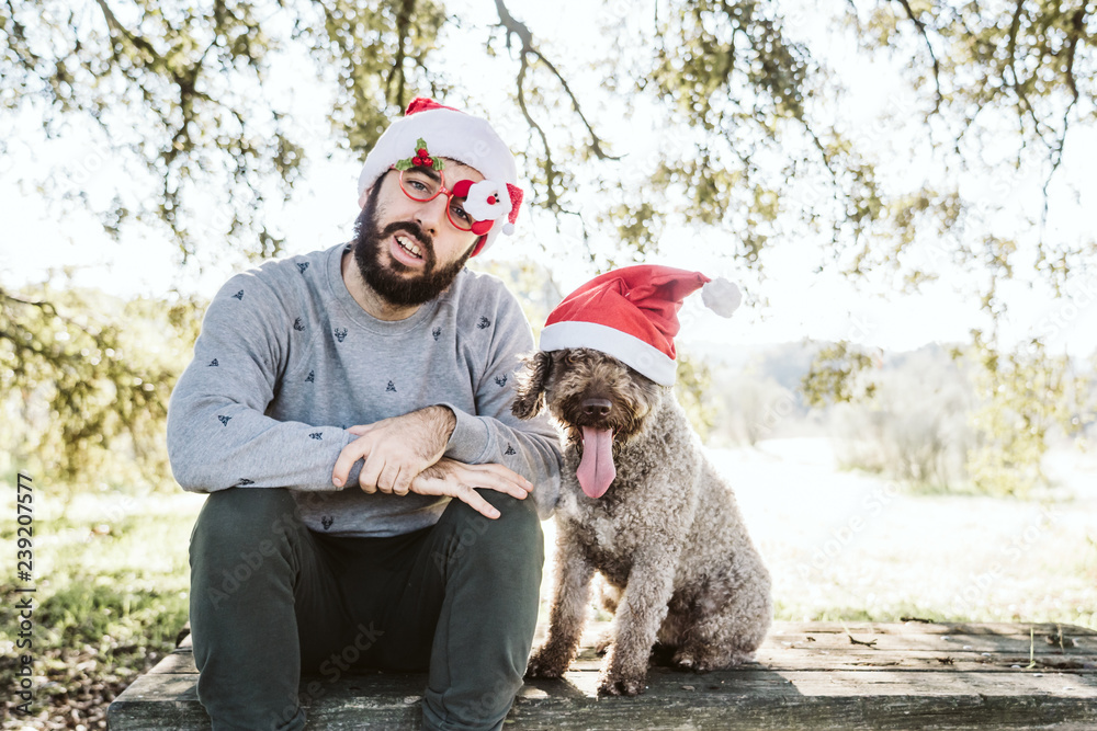 .Young man with his dog friend, both wearing santa claus caps celebrating Christmas on a sunny December day. Lifestyle