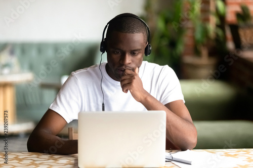 Focused African American man wear headphones, using laptop, looking at computer screen, thoughtful black student watching video, webinar, learning languages, studying online, making video call