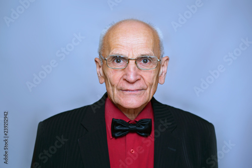 portrait of a senior elderly man in a red shirt, black jacket with a strip and a black bow tie seriously looking through glasses