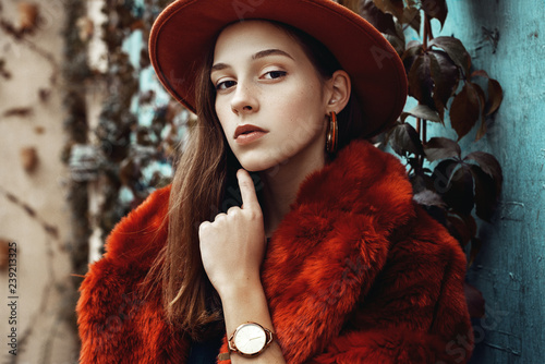 Εκτύπωση καμβά Outdoor close up fashion portrait of young beautiful confident woman wearing trendy orange faux fur coat, hat, hoop earrings, wrist watch, posing in street of european city