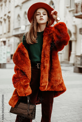 Outdoor fashion portrait of young beautiful confident woman wearing trendy orange faux fur coat, hat, green sweater, corduroy trousers, holding stylish handbag, posing in street of european city