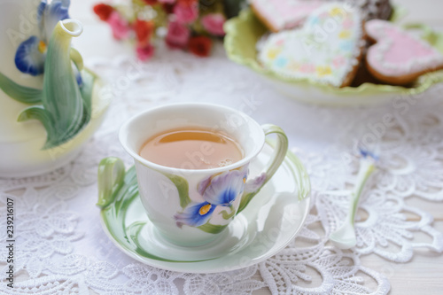  Beautiful porcelain cup with hot tea on a saucer next to nice teapot  against the background of a bouquet of flowers and a plate with heart-shaped gingerbreads on Valentine's Day.