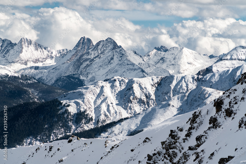 Fototapeta premium Snow covered Rocky Mountain ranges rise into while fluffy clouded skies