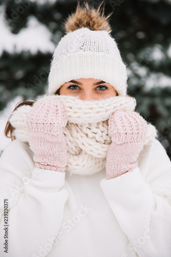 Outdoor close up portrait of young beautiful happy smiling girl wearing white knitted beanie hat, scarf and gloves. Model posing in park with Christmas lights. Winter holidays concept.