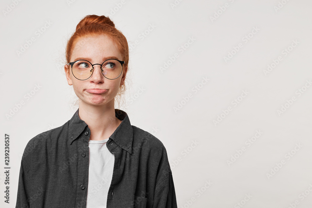 Portrait of a red-haired girl looks away wearing a men's black shirt and glasses expresses displeasure, sticks out her lower lip, empty copy space, white background Portrait of a red-haired girl looks away wearing a men's black shirt and glasses expresses displeasure, sticks out her lower lip, empty copy space, white background