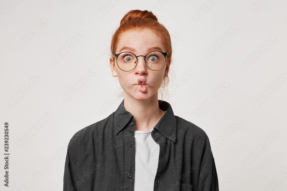 Red-haired girl portrays a fish with her lips looking through glasses with wide eyes. Woman doing fish face isolated on white background