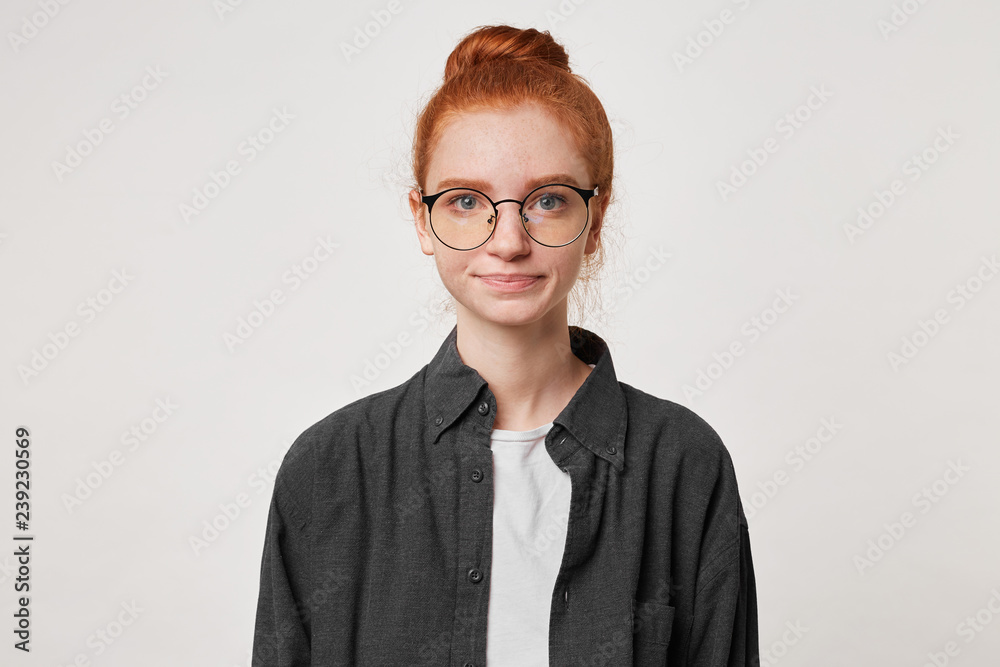 Portrait of calm confident girl with red hair gathered in a bun looks straight into the camera dressed in a black men's shirt isolated on a white background
