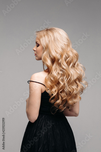 Back view of unrecognizable woman in black with volumous, blonde wavy hair over grey background. Isolated on grey. Studio. Perfect healthy hair. Hairdresser. photo