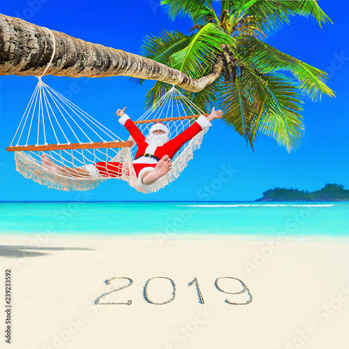Positive Christmas Santa Claus relax in hammock at palm beach and Happy New Year 2019 caption at white sand