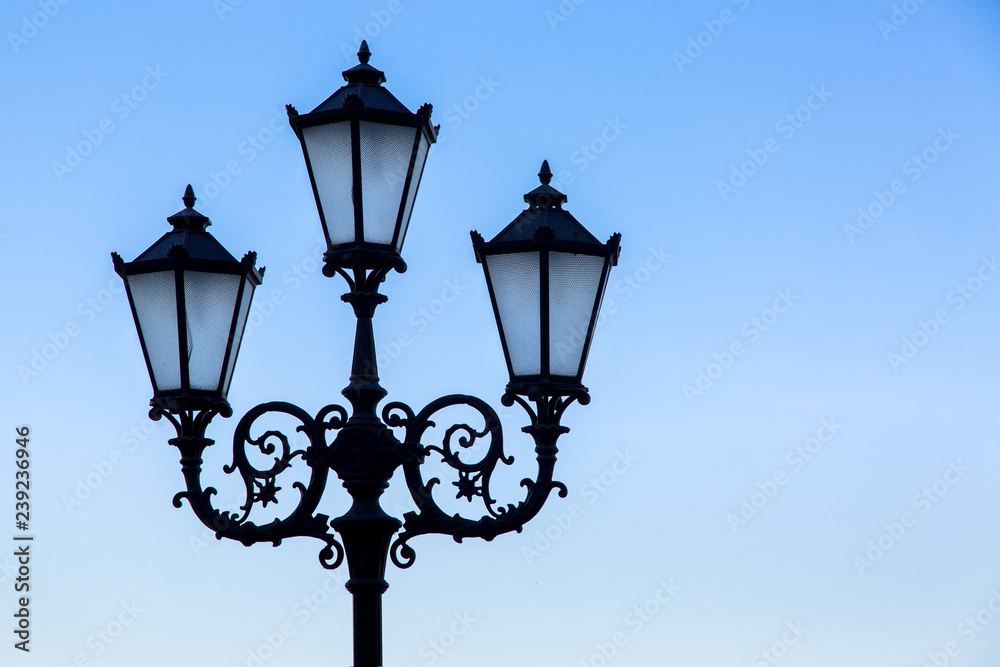 Silhouette of a black street lamp against a blue sky, lamppost with three wrought iron lamps with glass inserts.