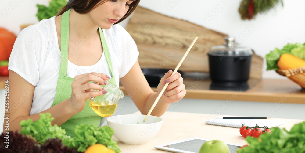 Beautiful Hispanic woman cooking in kitchen while using tablet computer and wooden spoon. Housewife found new recipe for dinner or breakfast. Healthy meal and householding concepts Beautiful Hispanic woman cooking in kitchen while using tablet computer and wooden spoon. Housewife found new recipe for dinner or breakfast. Healthy meal and householding concepts