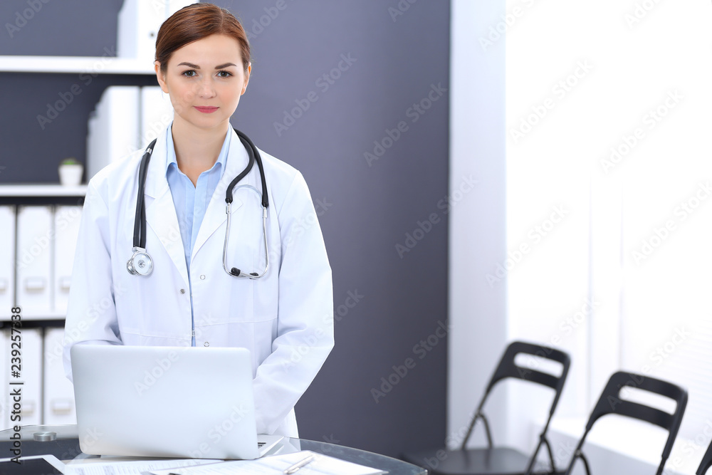 Happy doctor woman at work. Portrait of female physician using laptop computer while standing near reception desk at clinic or emergency hospital. Medicine concept Happy doctor woman at work. Portrait of female physician using laptop computer while standing near reception desk at clinic or emergency hospital. Medicine concept