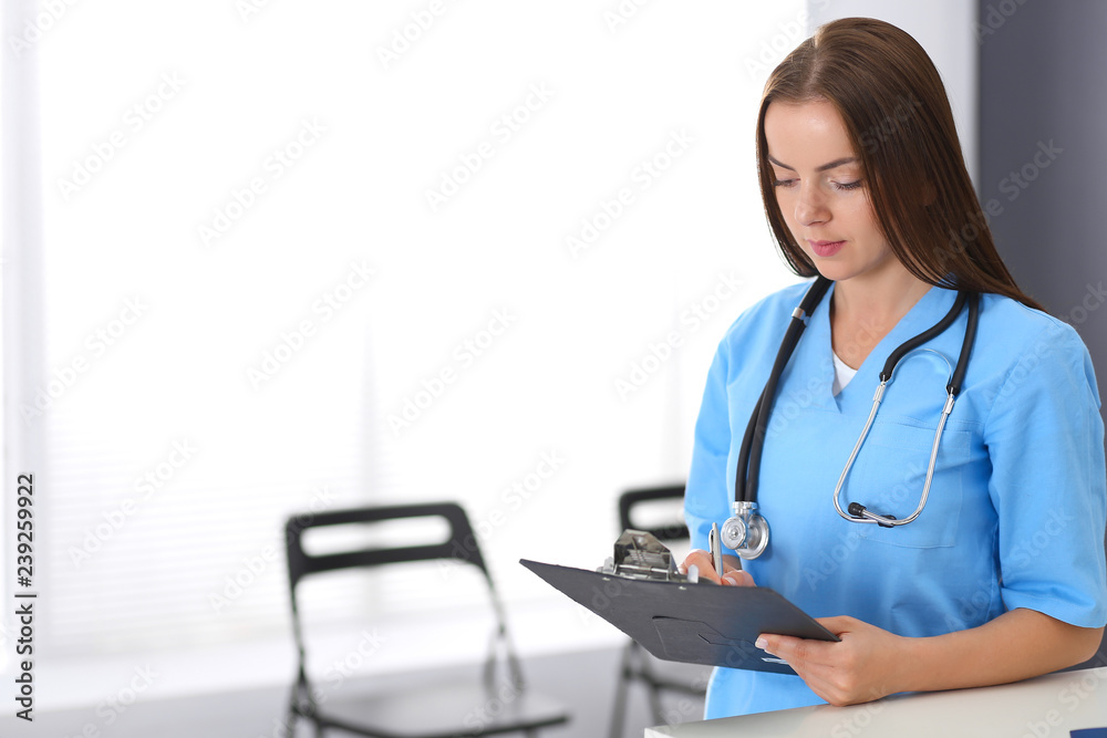 Doctor woman at work. Portrait of female physician filling up medical form while standing near reception desk at clinic or emergency hospital. Medicine and healthcare concept Doctor woman at work. Portrait of female physician filling up medical form while standing near reception desk at clinic or emergency hospital. Medicine and healthcare concept