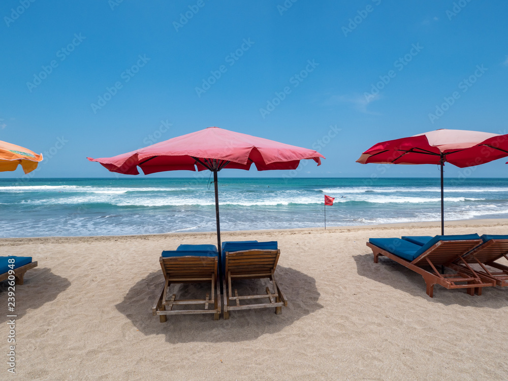 Red sun umbrellas stay on white sand beach with blue sea and blue sky on background. Concept for rest, relaxation, holidays, spa, resort. Bali, Indonesia, october 2018
