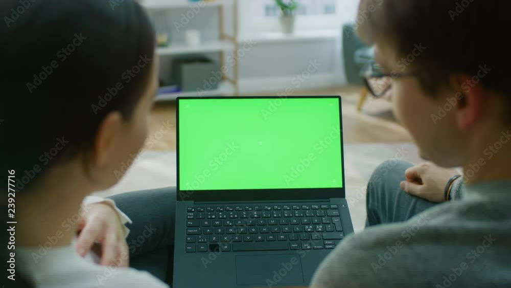 Young Man and Woman at Home Using Green Mock-up Screen Laptop Computer While Sitting on Couch in Living Room. A Couple in Love Doing Internet Shopping. Zoom out Back View Camera Shot. Young Man and Woman at Home Using Green Mock-up Screen Laptop Computer While Sitting on Couch in Living Room. A Couple in Love Doing Internet Shopping. Zoom out Back View Camera Shot.
