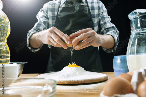 Professional male cook sprinkles dough with flour, preapares or bakes bread or pasta at kitchen table, has dirty uniform, isolated over black chalk background. Baking concept photo