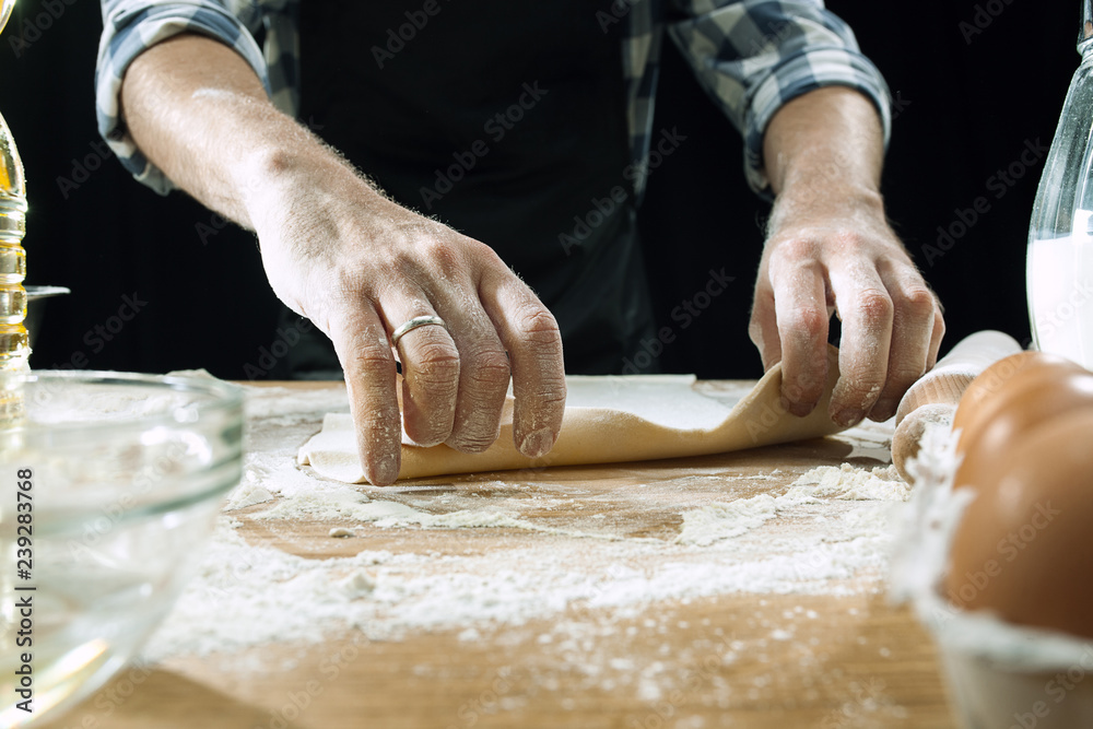 Professional male cook sprinkles dough with flour, preapares or bakes bread or pasta at kitchen table, has dirty uniform, isolated over black chalk background. Baking concept