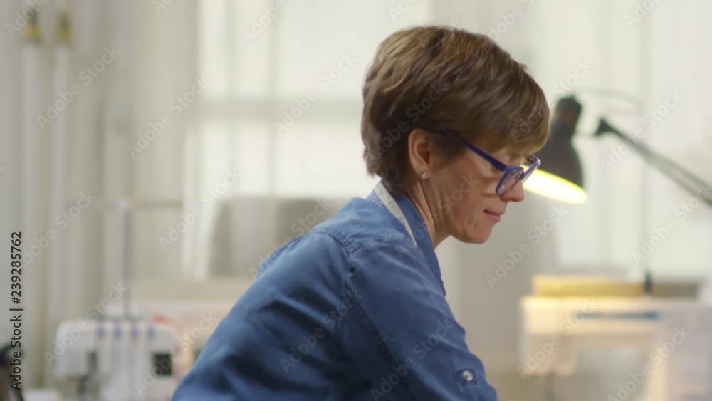 Tilt down of experienced Caucasian seamstress in eyeglasses making marks tailors chalk on fabric and using ruler while working in sewing studio Tilt down of experienced Caucasian seamstress in eyeglasses making marks tailors chalk on fabric and using ruler while working in sewing studio