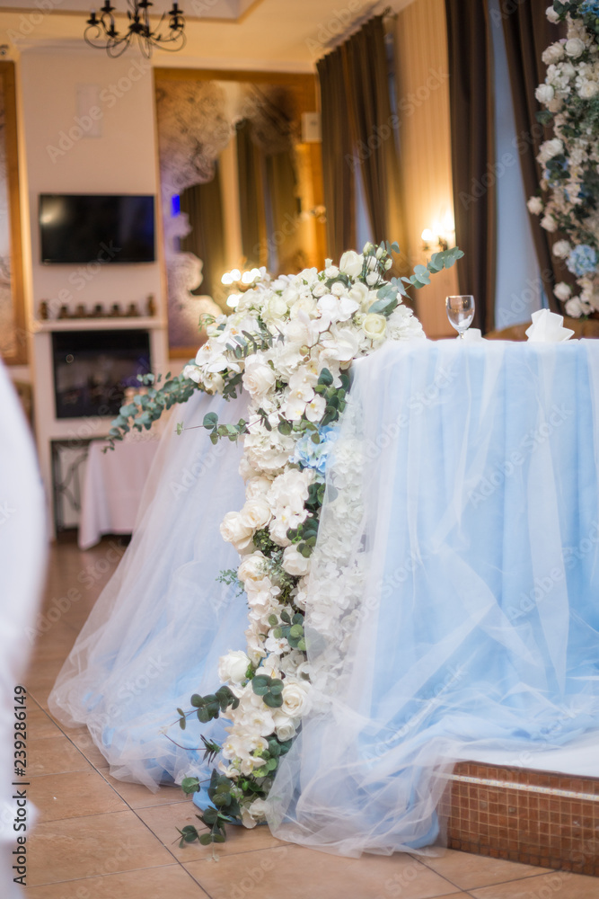 Wedding presidium in restaurant, free space. Wedding banquet table for newlyweds with flowers, greenery, pink cloth and bulbs. Lush floral arrangement on wedding table. Luxury wedding decorations