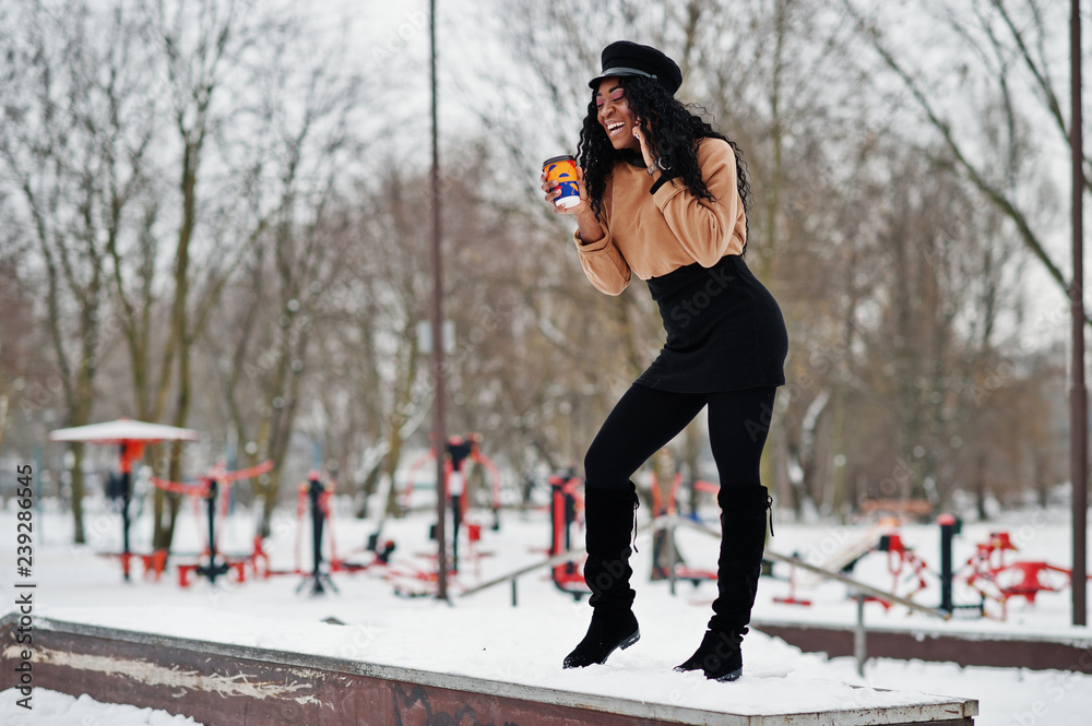 African american woman in black skirt, brown sweater and cap posed at  winter day against snowy background, holding cup of coffee and speaking on  phone. Stock Photo | Adobe Stock