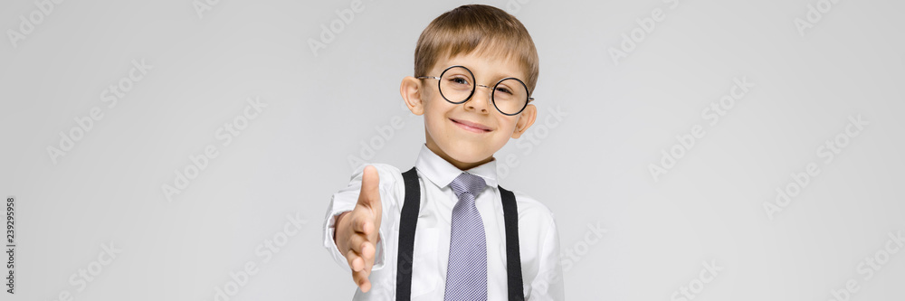 A charming boy in a white shirt, suspenders, a tie and light jeans stands on a gray background. The boy stretches out his hand forward